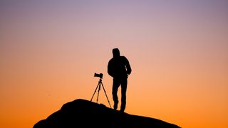 Silhouette of man with one of best low-light cameras on tripod, standing on hillside