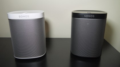 Två stycken Sonos Play:1, en vitgrå och en svartgrå, står bredvid varandra på en svart yta mot en vit vägg.