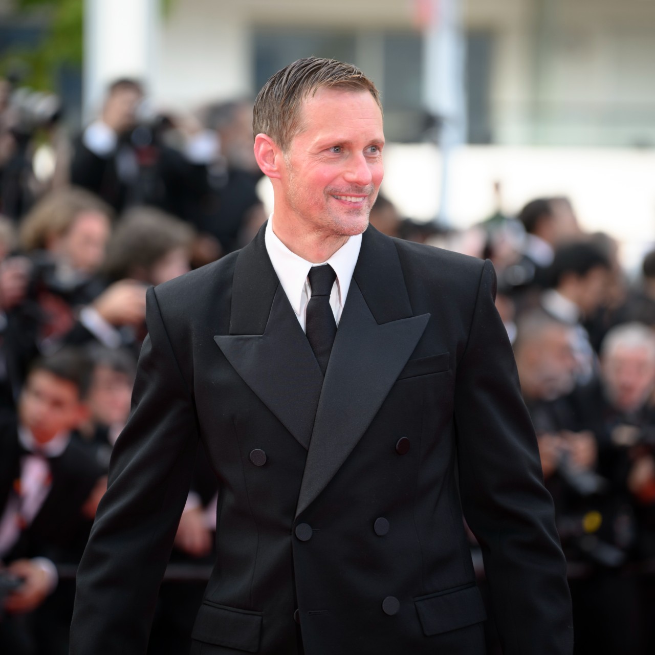 a white man wearing a black tuxedo posing in front of paparazzi on a red carpet 