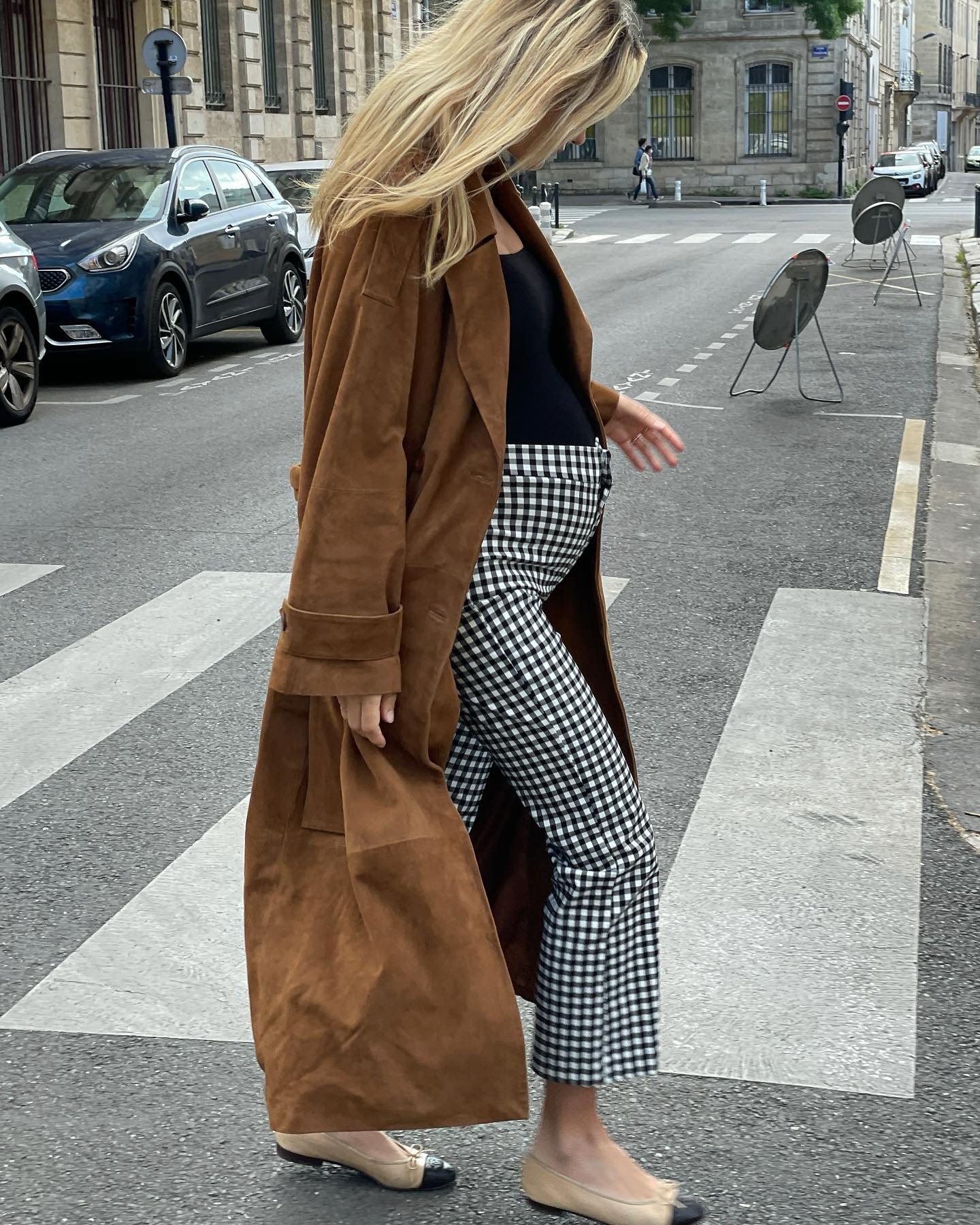 French influencer Anne-Laure Mais walks across a Paris street wearing a chic maternity outfit with a long suede coat, a black top, cropped gingham-print pants, and two-tone Chanel ballet flats