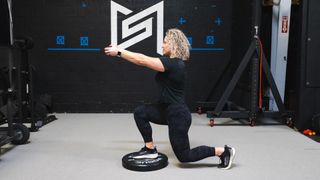 Woman stands in gym, with one foot on a weight plate on the floor and the other behind her, both knees bent to 90° and with her arms extended in front of her. She wears a black T-shirt, black leggings and black trainers.