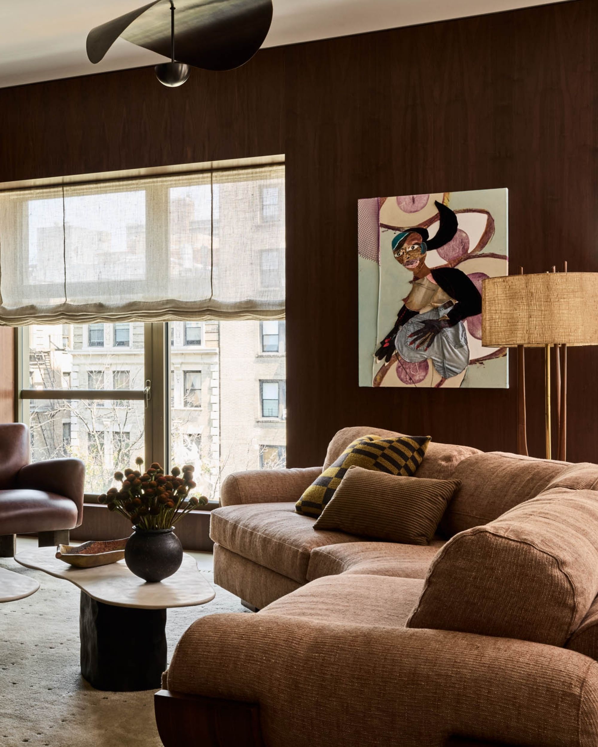 A cozy living room with dark brown wooden walls and a large, caramel brown plush, curved sofa. There is a window on the back wall with sheer roman blinds and there is a floor lamp behind the sofa.