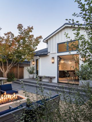 a backyard of a home with trees and plants placed all around. The exterior of the home is black and grey and you can see inside the bright and chic home.