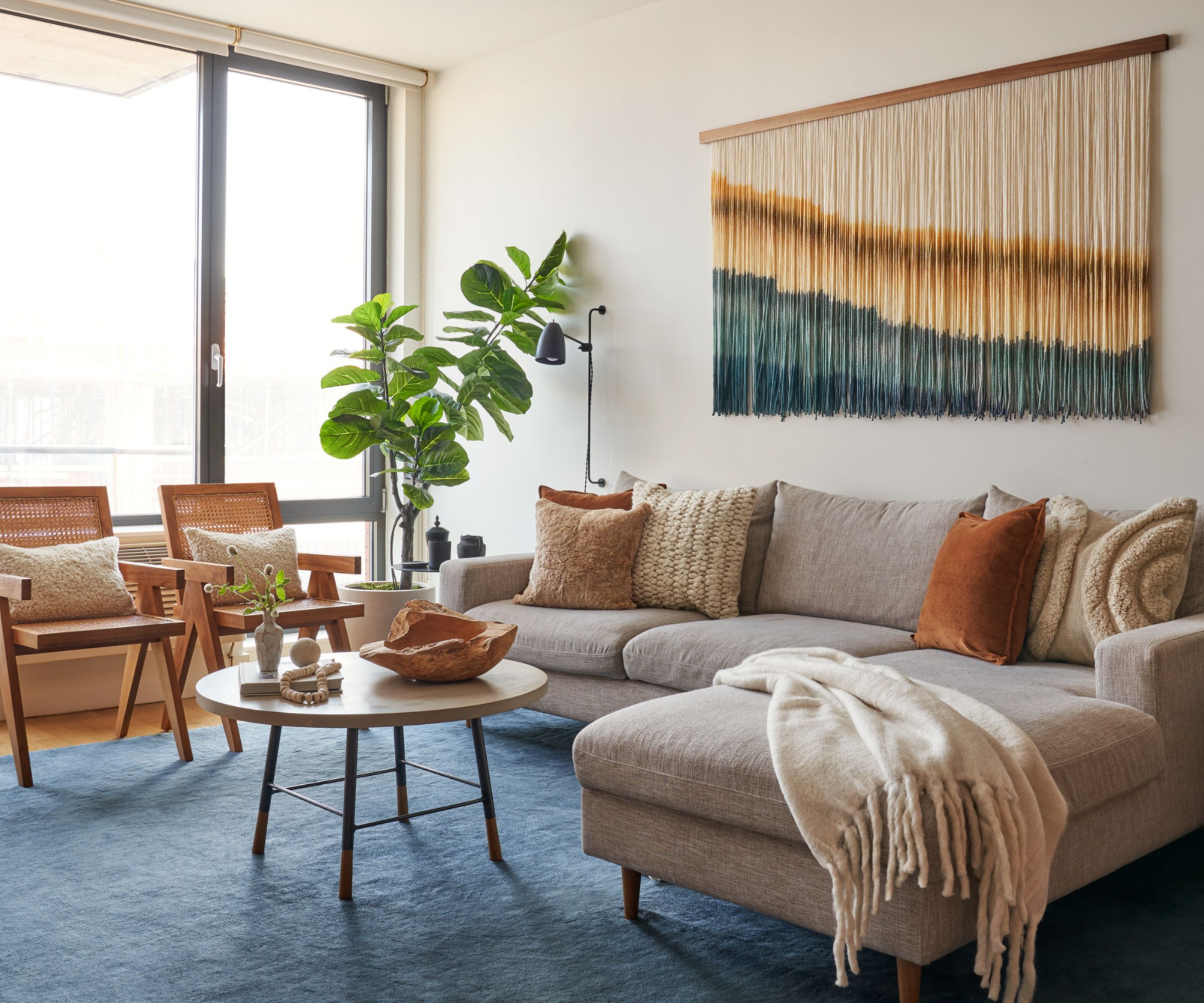 neutral apartment living room with a neutral corner sofa and touches of orange and blue with a large blue rug, orange cushions, and a fringed orange and blue wall hanging