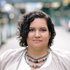 A photo of Karen Ortiz standing on a city street, wearing a tan jacket and layered beaded necklaces, with a calm, determined expression.