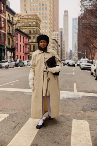 A New York Fashion Week guest wearing a tan trench coat, tan skirt, tan top, white tights, black shoes, black fuzzy hat