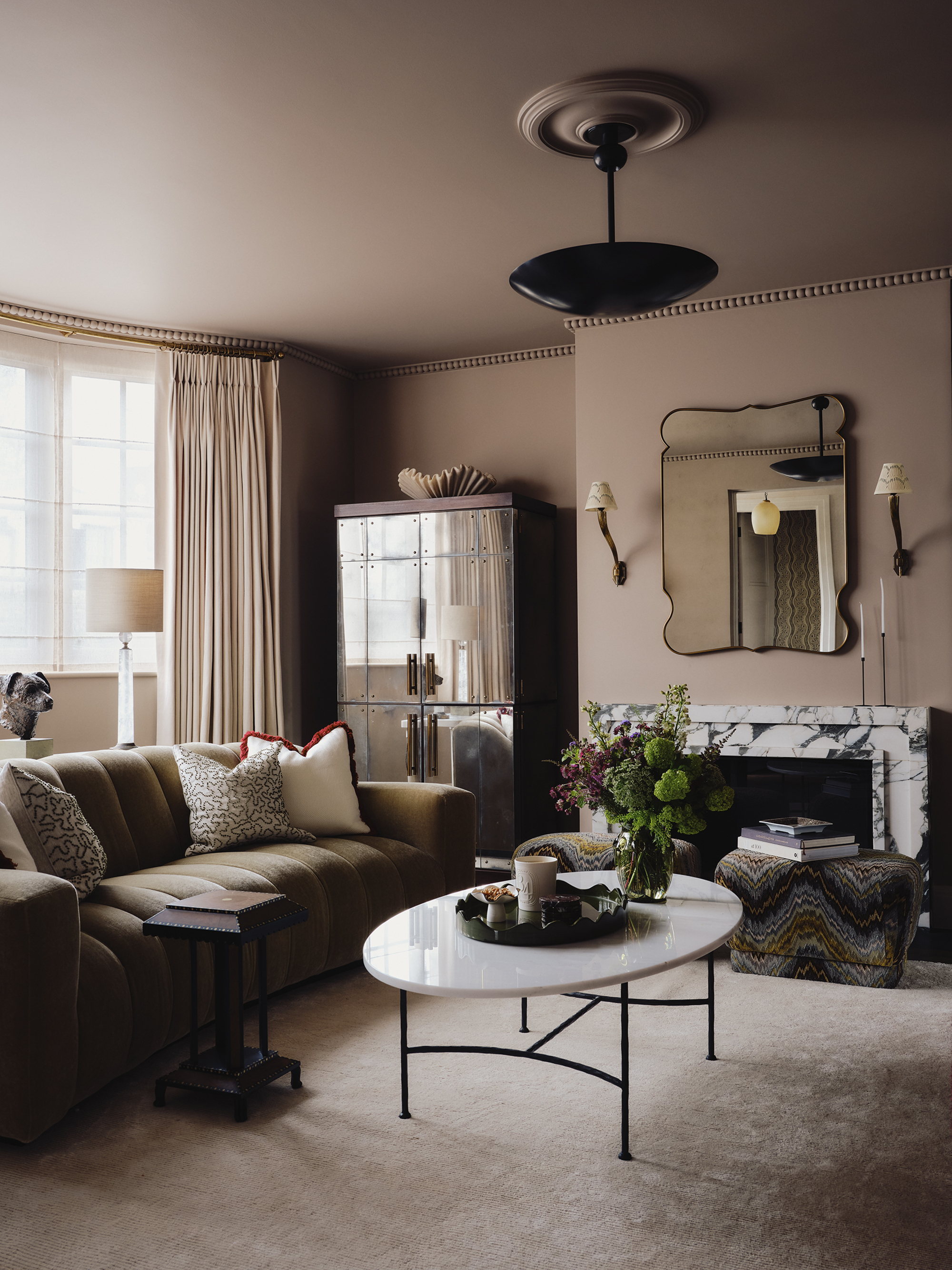 cozy living room with taupe color-drenched walls, white curtains, marble fireplace with mirror hanging above with wall sconces, mirrored cabinet, tan sofa with cushions in animal print, white marble coffee table with black legs and beige rug