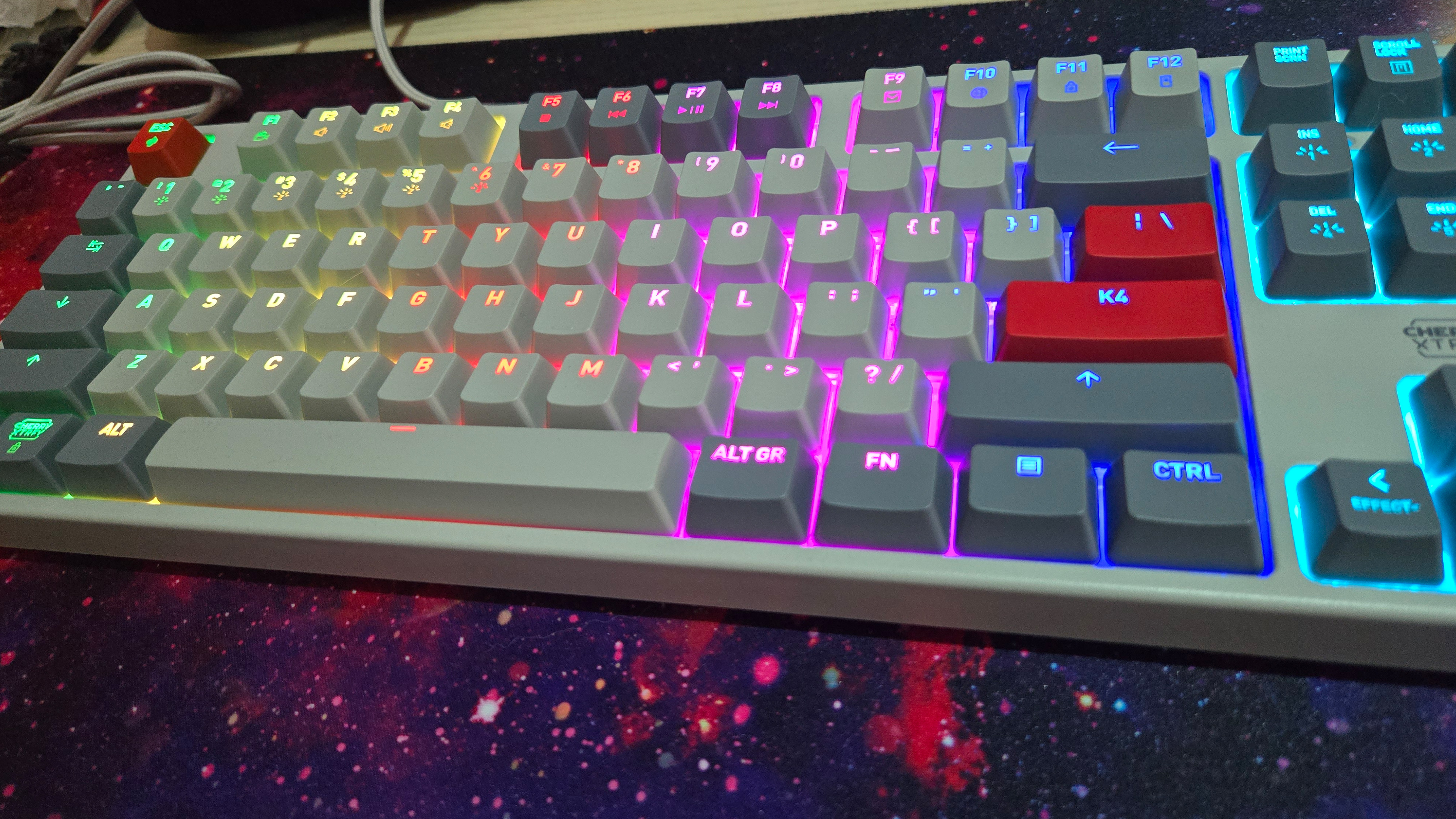 A closer look at the Cherry Xtrfy K4V2 TKL in grey with red accent keys. It sits on a large mousepad showing a nebula. The RGB lights are on.