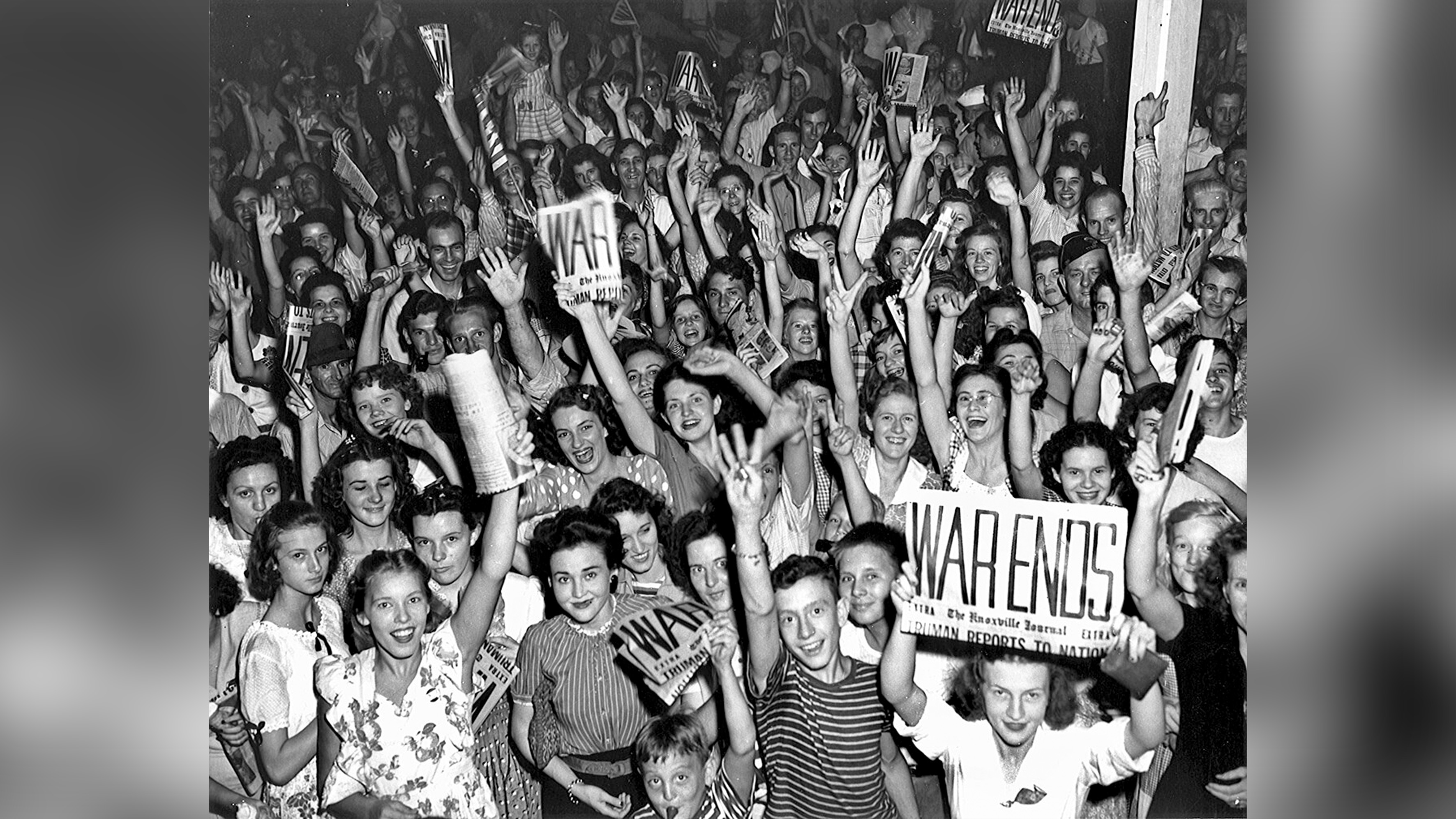 A crowd celebrating. Some people are holding newspapers with the headline "WAR ENDS".
