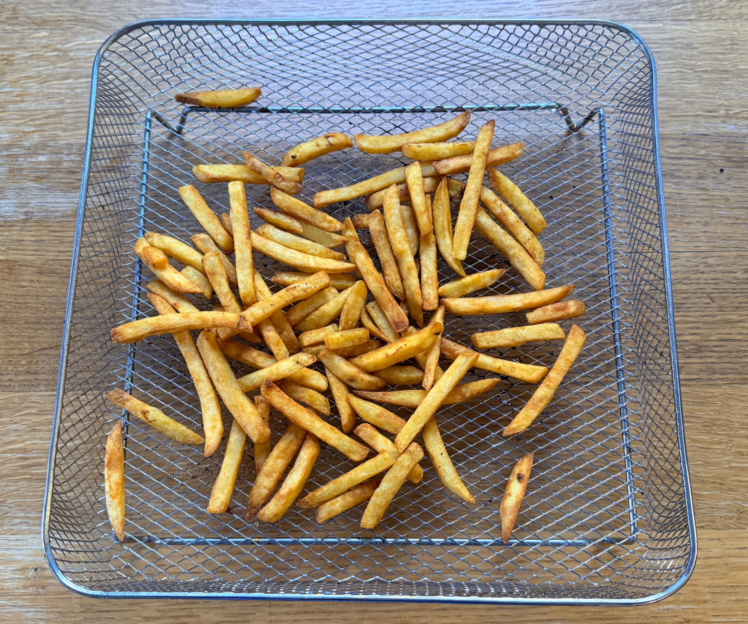 Thin cut French fries baked in a wire mesh basket on a wooden kitchen counter.