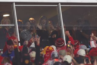 Taylor Swift waves to the crowd from her box seats at the Chiefs game wearing a black long-sleeve shirt with high waist shorts