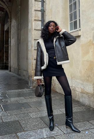 A black turtleneck outfit is shown in a photo of a French woman on the sidewalk wearing a black shearling leather jacket over a black turtleneck with a black mini skirt, designer belt, black tights, black knee-high boots, and a small bag