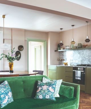 A pale pink-painted open-plan kitchen with a living room with a green velvet sofa