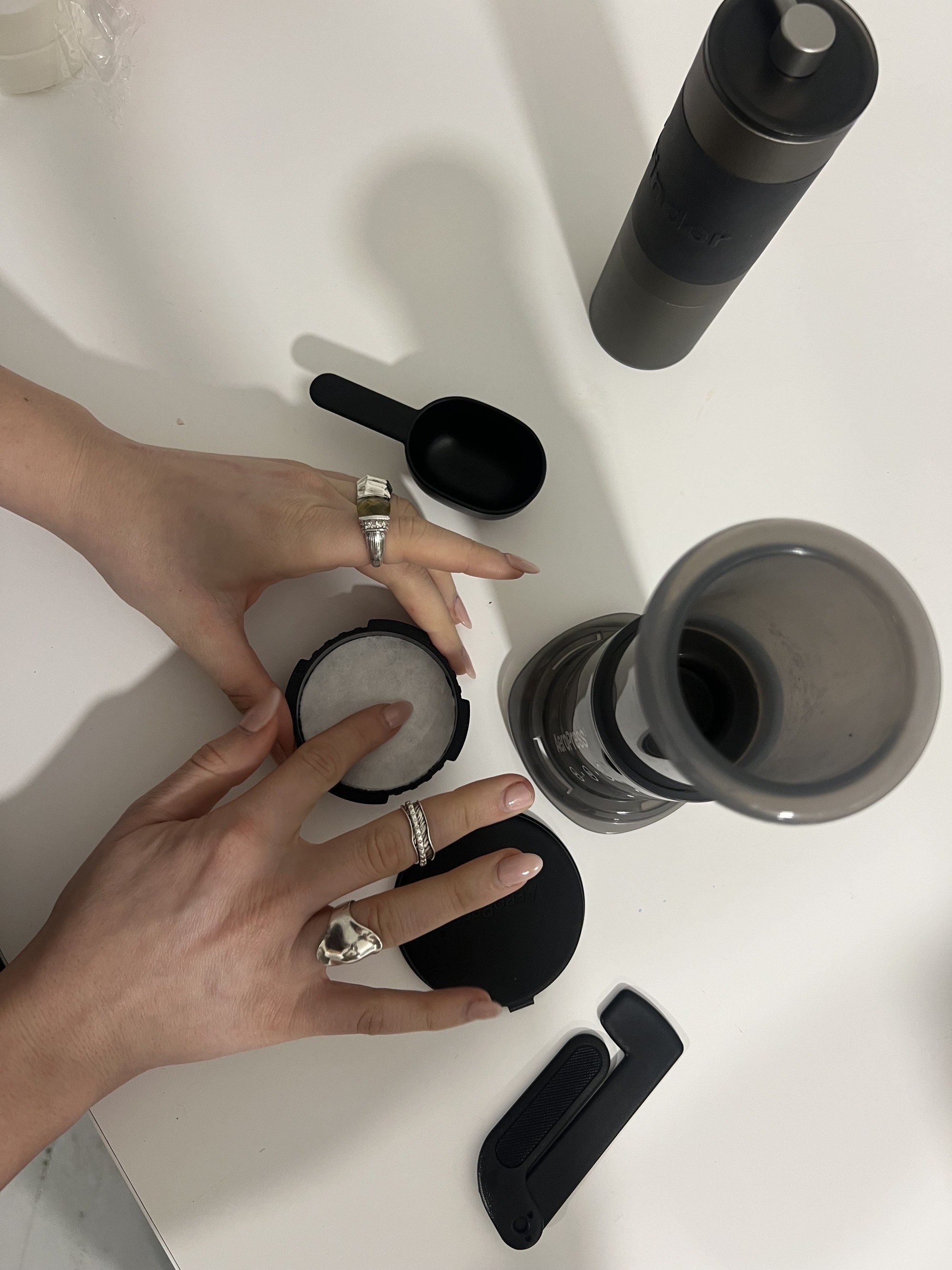 Image of two hands from above placing a coffee filter into the top of the AeroPress coffee maker on a white countertop. There are other black coffee making accessories on the counter.