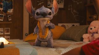 Still from the live-action movie version of Lilo & Stitch. A blue, fluffy, koala-like alien is standing on a bed and is wearing a yellow Hawaiian lei necklace around his neck.