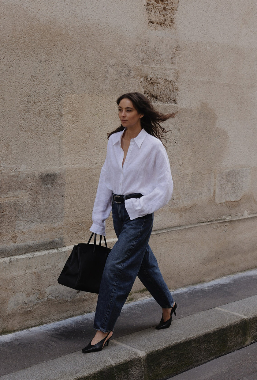 A woman walking down the sidewalk wearing a white button-down shirt tucked into dark wash barrel leg jeans styled with a black belt, black pointed slingback heels, and a black tote bag.