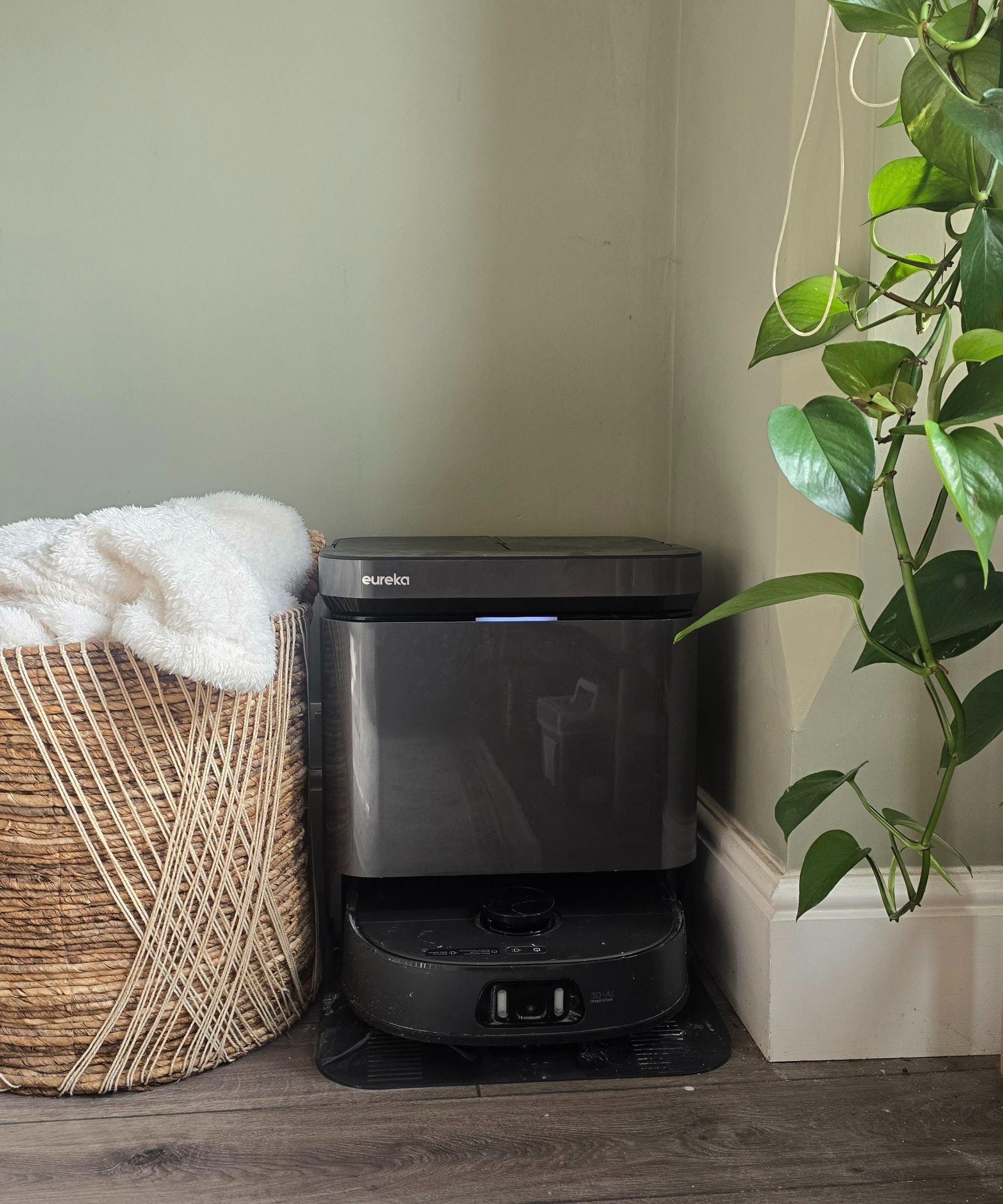 A black Eureka J15 Pro Ultra robot vacuum next to a woven basket and a leafy green plant
