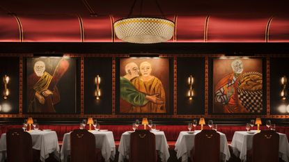 A dimly lit, theatrically furnished restaurant dining room features large figurative paintings depicting old men in togas against red backgrounds, with spotlights on each of them, traditionally set tables with red velvet banquettes and chairs, and golden table lamps and lighting.