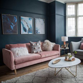a living room with dark blue walls, pink velvet sofa with fluffy cushions, a marble and gold coffee table and three framed artistic blue and gold pictures on the wall