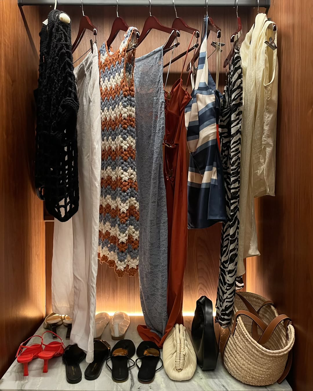 A hotel room wardrobe with clothes unpacked on the hangers and shoes and handbags arranged on the shelf.