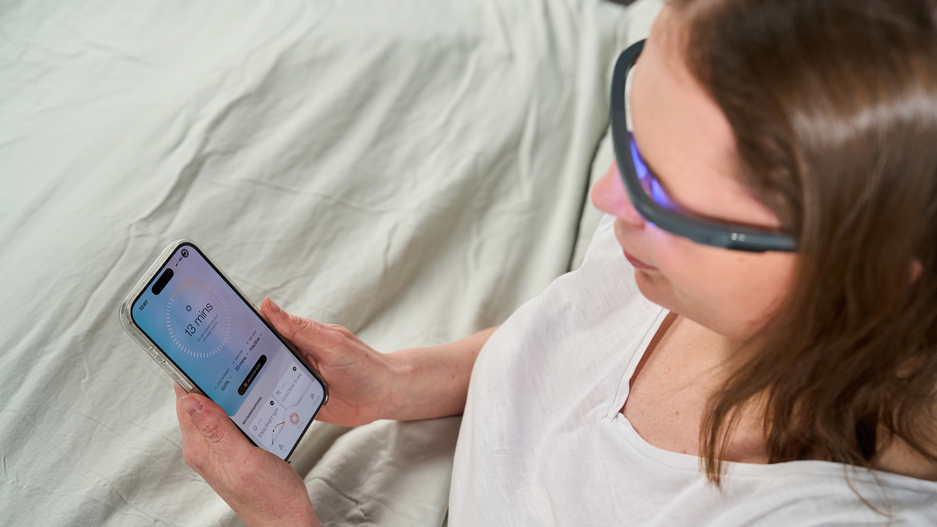 A woman with brown hair sits up in bed while wearing the AYO+ Light Therapy Glasses straight after waking