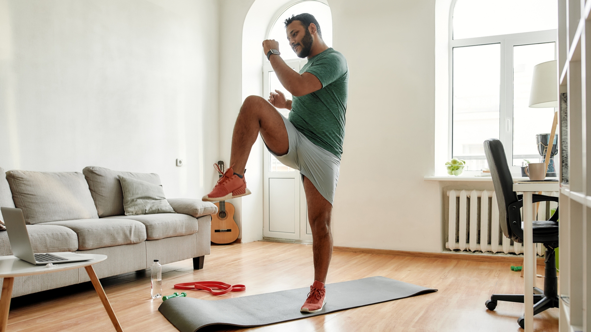 man wearing tshirt and shorts exercising in a living room setting standing on an exercise mat performing high knee to opposite elbow move. 