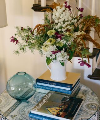 A table in an entryway with stacks of books, a floral arrangement and a white and blue tablecloth