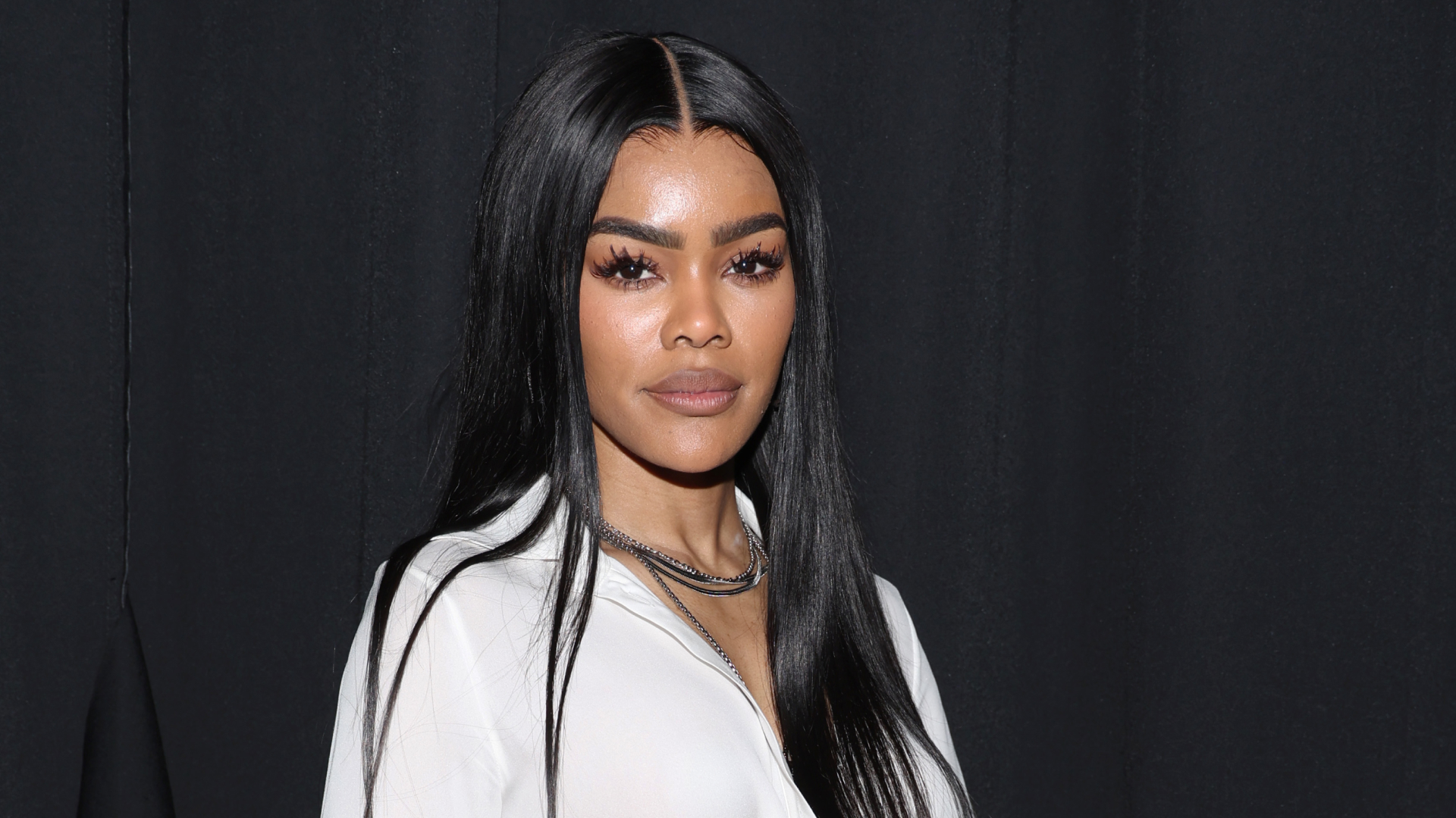 Teyanna Taylor standing in front of a black backdrop wearing a white shirt and necklace. She has long straight hair. 