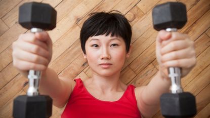 Woman lying on wooden floor holding dumbbells above her