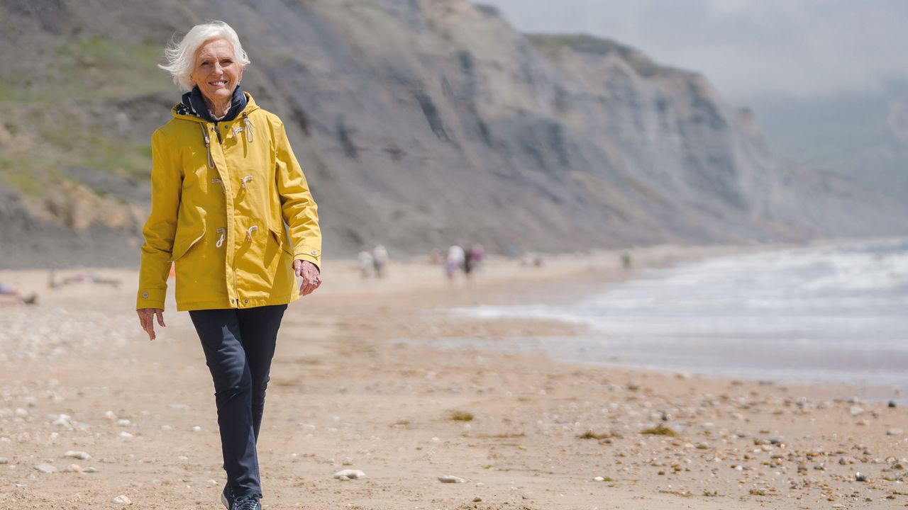 Mary Berry walking along a beachfront wearing a yellow raincoat