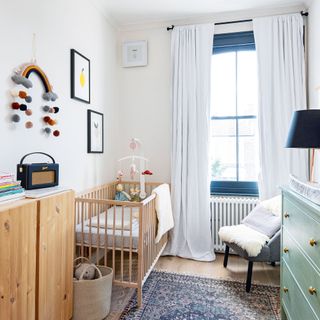 White nursery with wooden cot, blue window frames, white blackout curtains, blue Persian rug, rainbow wall hanging, mobile