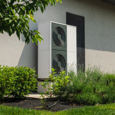 Heat pump installed on the outside of a rendered house, surrounded by greenery in the garden