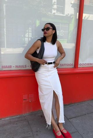 A denim skirt outfit is shown in a photo of a woman leaning against a storefront wearing a white tank top with a black belt, a white denim maxi skirt, a black woven shoulder bag, and red mesh ballet flats