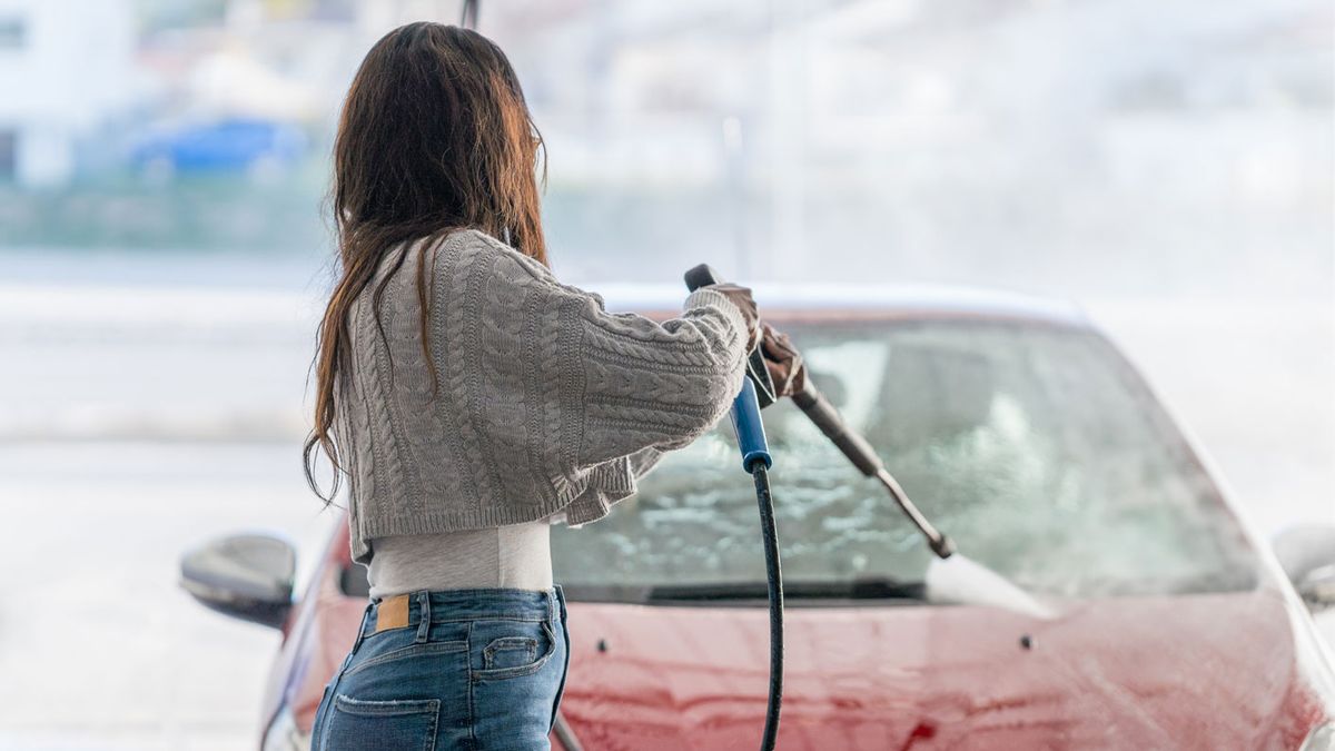 How to wash a car with a pressure washer: Image shows a woman in a grey jumper using a pressure washer to wash her red car.