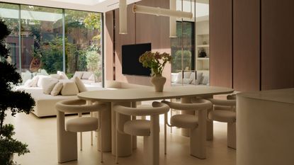 An oval-shaped cream dining table with cylindrical legs on either side, has cream curved back modern dining chairs, and abstract pendant lighting hanging above, along with large glass doors to outside