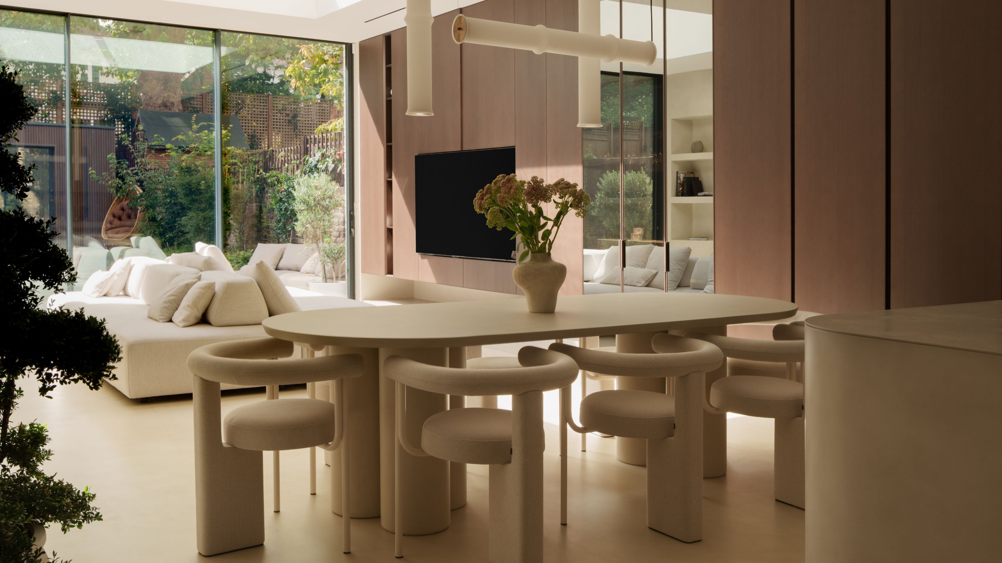 An oval-shaped cream dining table with cylindrical legs on either side, has cream curved back modern dining chairs, and abstract pendant lighting hanging above, along with large glass doors to outside