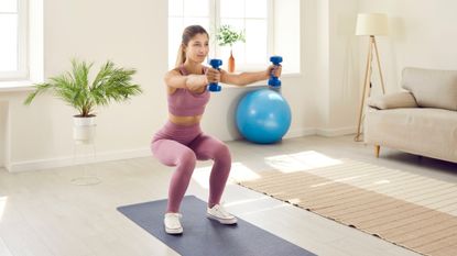 Woman squats while holding two light dumbbells arms extended in front living room setting wearing pink crop top leggings 