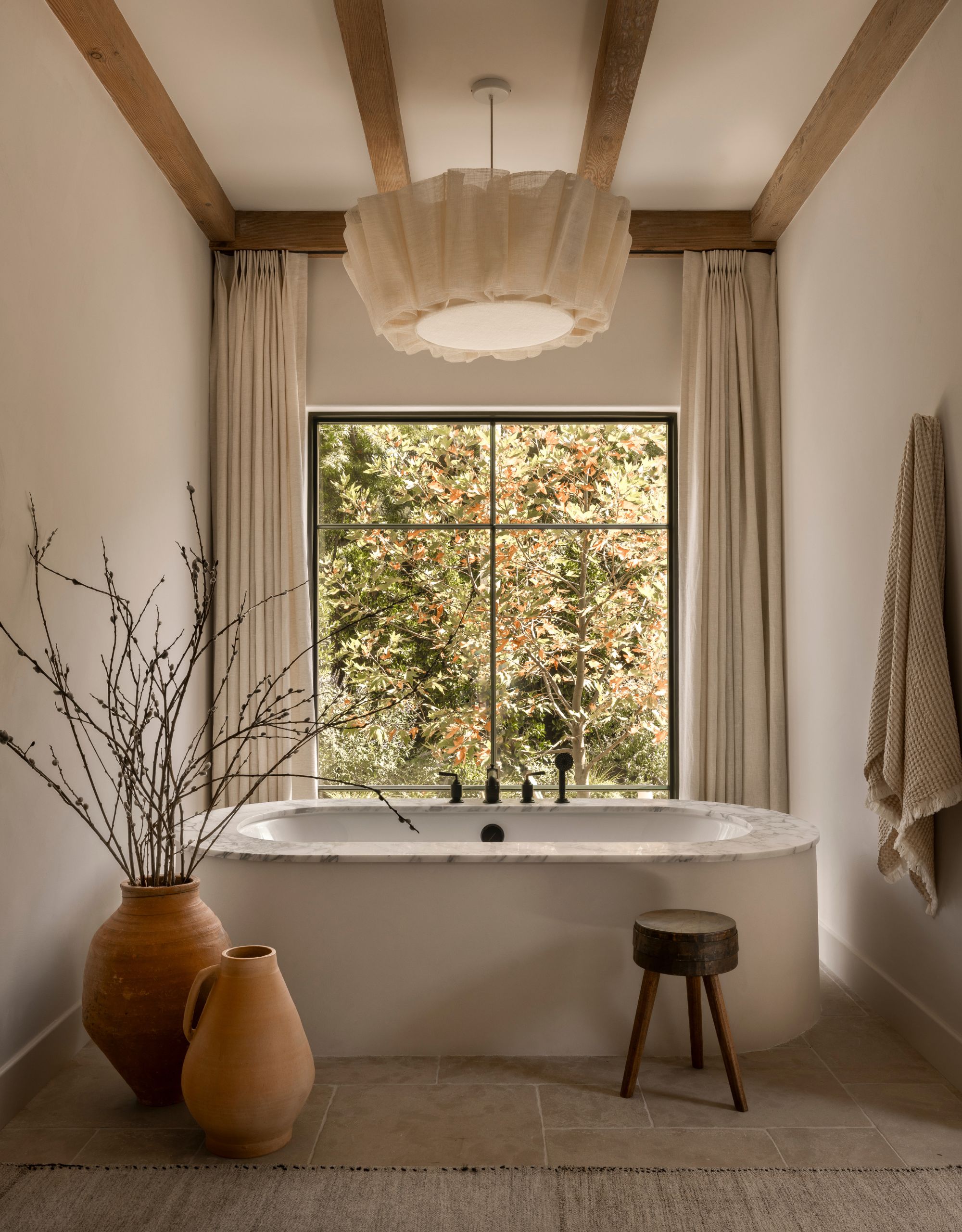 A modern neutral bathroom with a freestanding next to large vases, a delicate fabric pendant light, exposed beams on the ceiling and a view of the outdoors framed by floor-length cream curtains