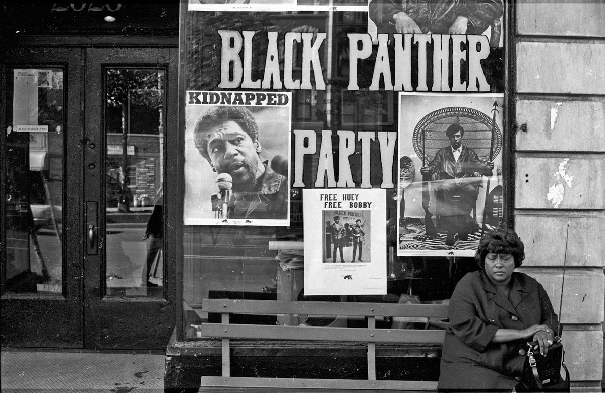 A woman sits on a bench outside the Black Panther office in Harlem circa 1970 in New York City, NY. Pictured in the window are Panther founders Huey P. Newton and Bobby Seale. 