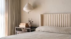 A minimalist bedroom with a pendant lamp, a nightstand with a vase and a set of burl wood boxes, near a large bed with a beige headboard and a minimalist bed