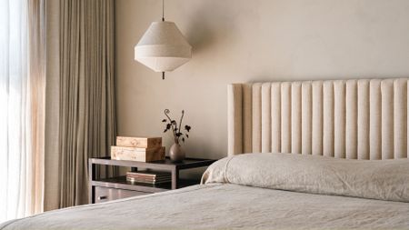 A minimalist bedroom with a pendant lamp, a nightstand with a vase and a set of burl wood boxes, near a large bed with a beige headboard and a minimalist bed