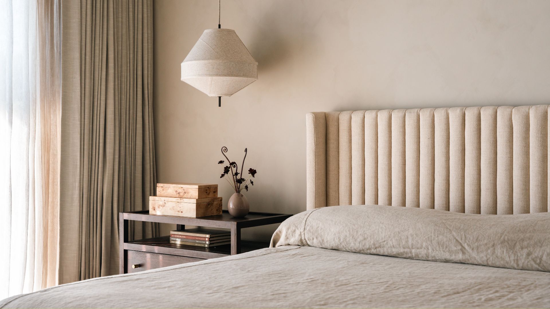 A minimalist bedroom with a pendant lamp, a nightstand with a vase and a set of burl wood boxes, near a large bed with a beige headboard and a minimalist bed