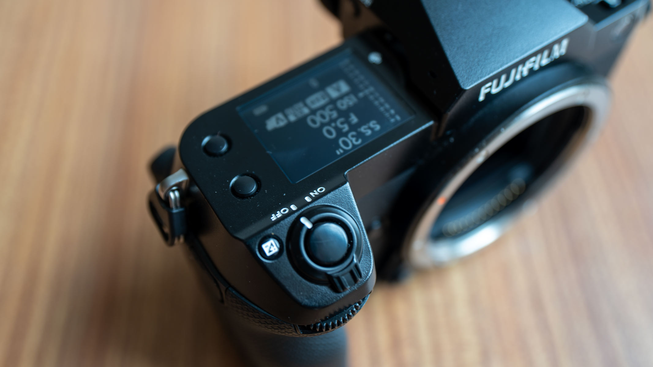 A close-up of the power button, exposure compensation button and secondary LCD monitor of the Fujifilm GFX100S II, on a wooden table.