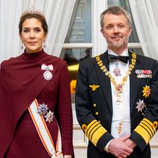 Queen Mary in a maroon gown and tiara standing with King Frederik in a military uniform and bow tie