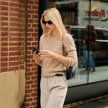 Woman in khaki trousers, beige short-sleeve sweater, sunglasses, and a chain-strap bag