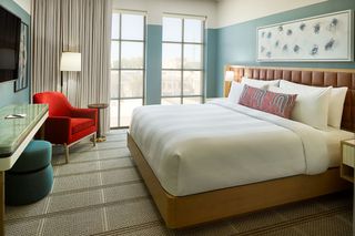 A luxury hotel bedroom features pastel shaded interior decor, upholstered furniture, a leather-like bedframe, and wide windows overlooking the city.