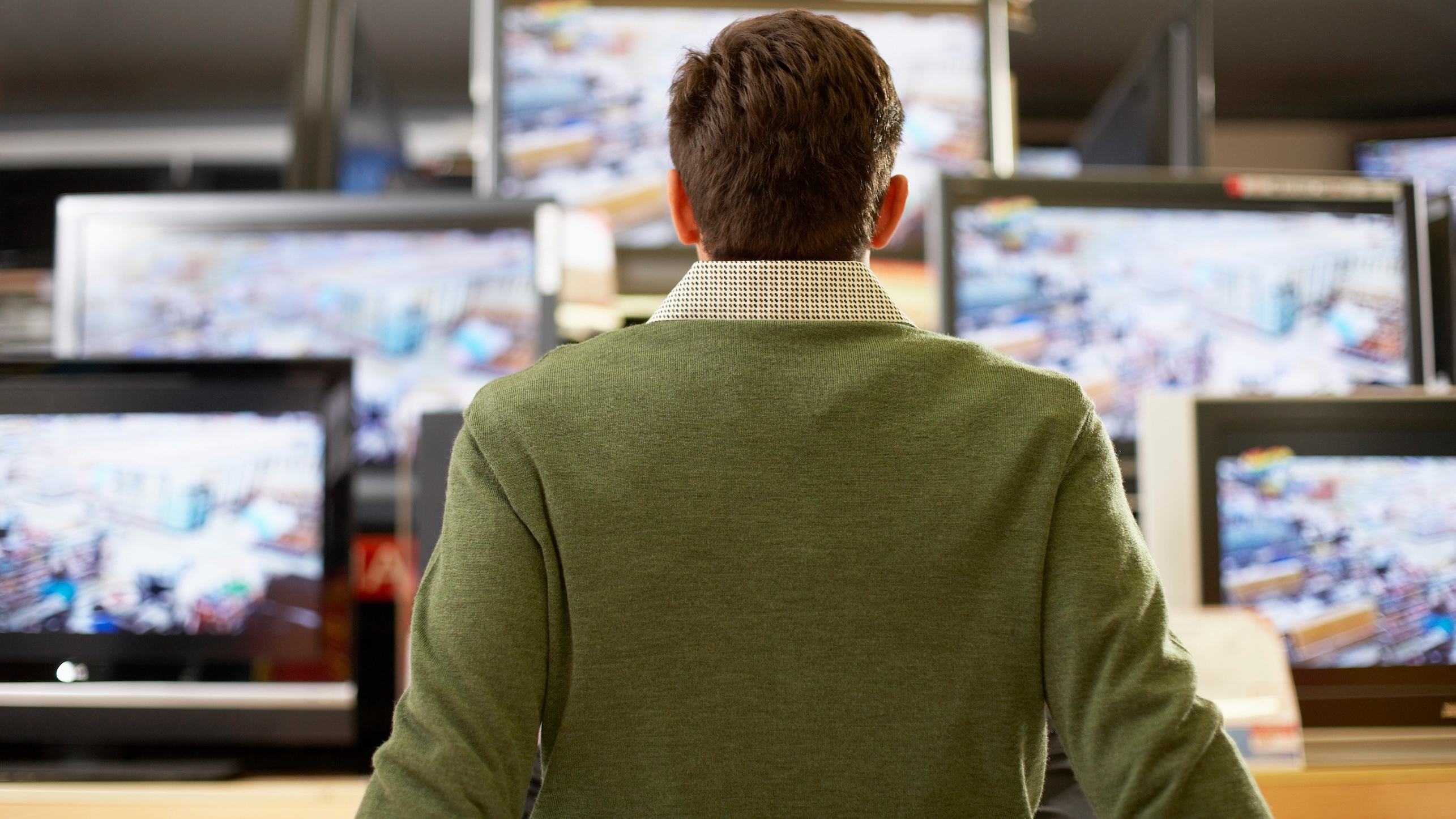 A man seen from behind sitting down in a shop, leaning back on his hands, taking in the rows of TVs in front of him.