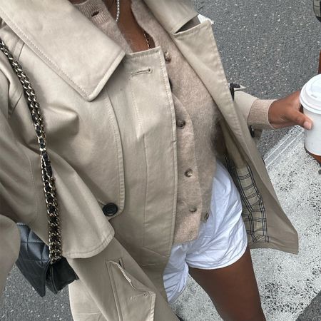 Claire Most wearing a tan trench coat, tan cardigan, and white shorts while walking and drinking of a coffee.