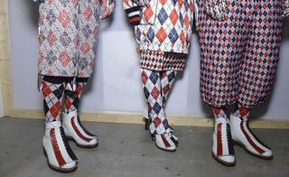 Three models wearing looks from Moncler Gamme Bleu's collection. They are wearing loose-fitting pants, a skirt and socks in red, blue and white with a repeating diamond pattern. Their shoes are white and feature red and blue stripes
