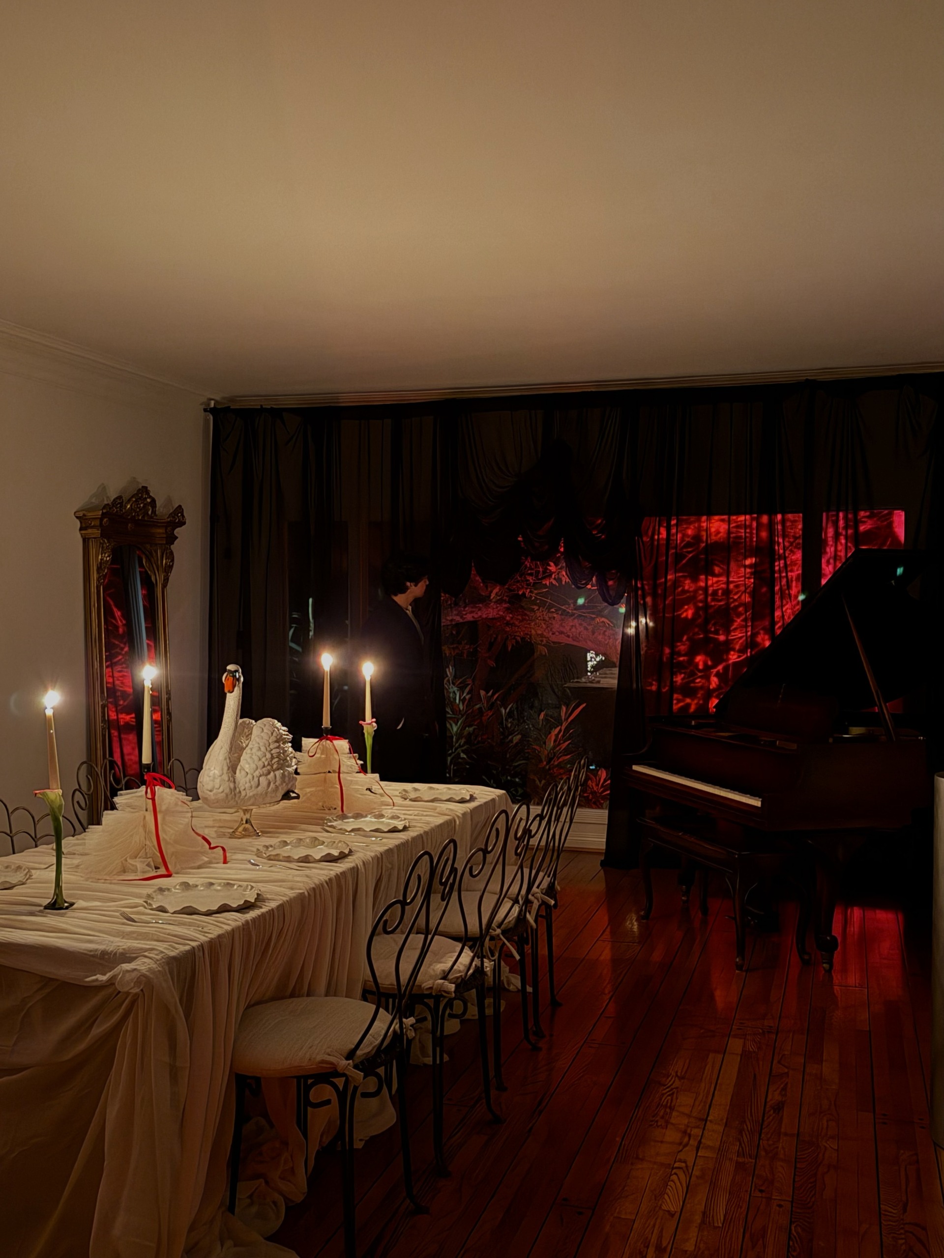 A dining area decorated for Halloween with black sheer curtains partly revealing a red-lit trees outdoors, a grand piano, and a table set with a swan statue in the center among taper candles